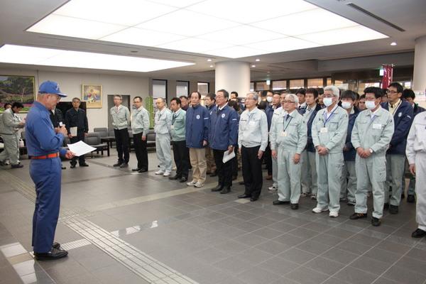 ロビーにて消防の帽子を被り消防服姿の市長がマイクを持って話をしているのを作業服姿の職員が整列して聞いている写真